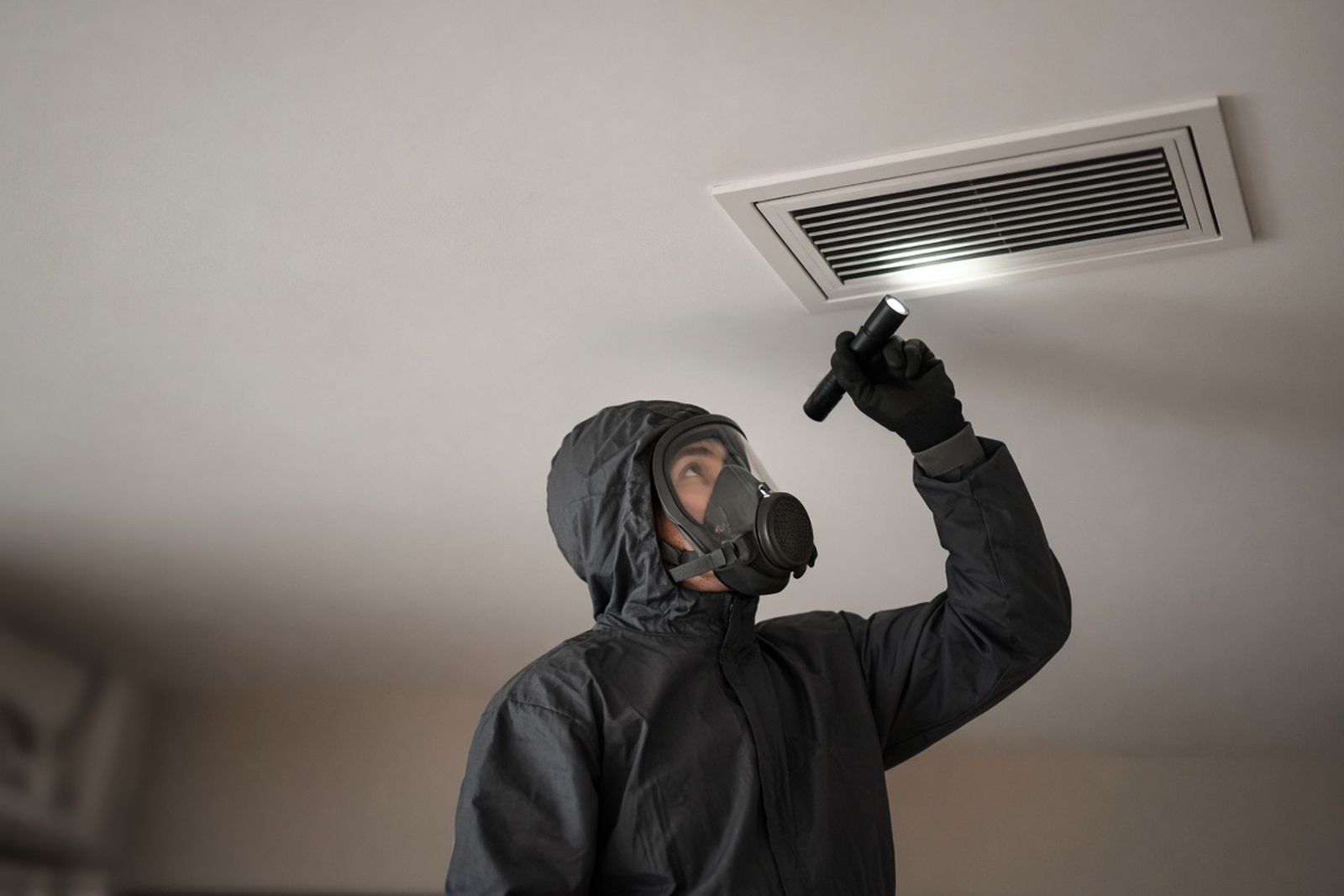 Technician in a respirator inspecting a ceiling air vent with a flashlight